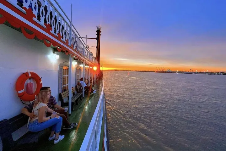 People on a boat enjoying a sunset over a calm river with a scenic city skyline in the distance.