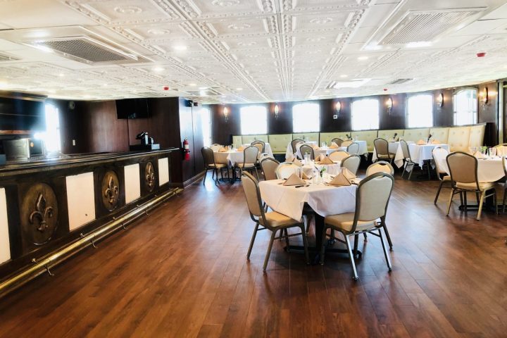 Elegant dining room with round tables and white tablecloths, wooden floor, ornate bar, and decorative ceiling.
