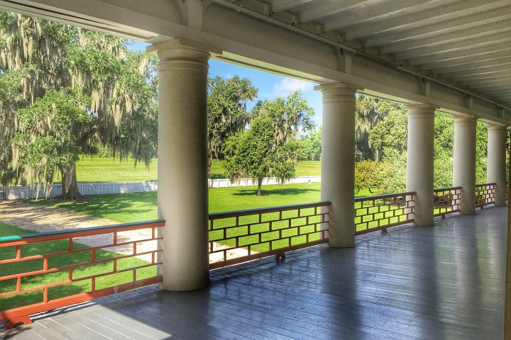 Veranda with columns overlooking lush lawn and trees on a sunny day.