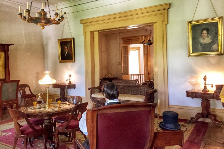 19th-century parlor with man in period attire, chandelier, portraits, and vintage furniture.