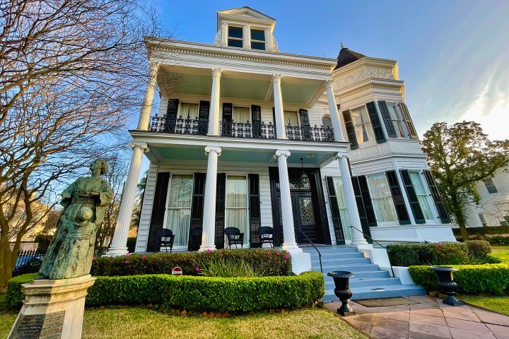 Victorian house with pillars, bay windows, and a garden statue in front under blue sky.