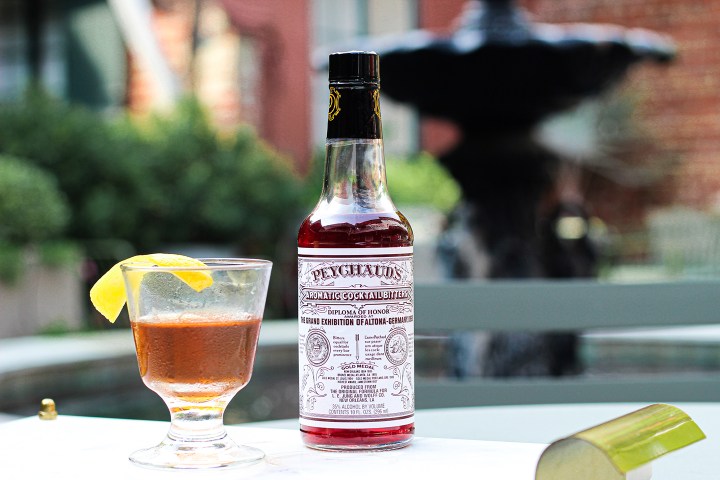 Bottle of Peychaud's bitters and cocktail on outdoor table near a fountain.