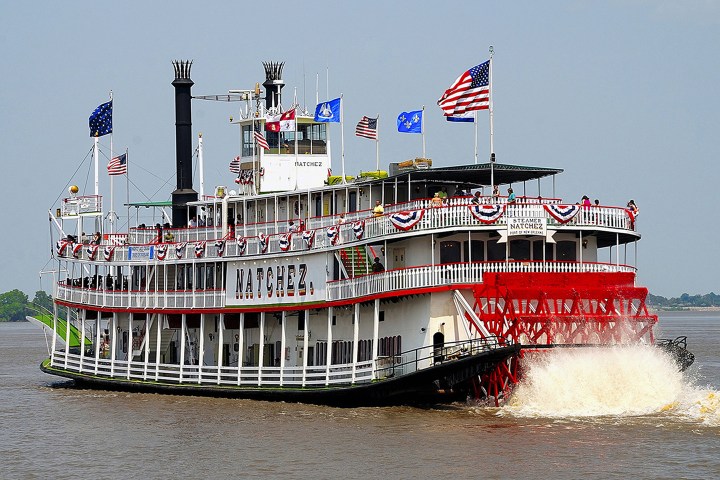 Paddle steamboat Natchez with flags sails on a river under a clear sky.