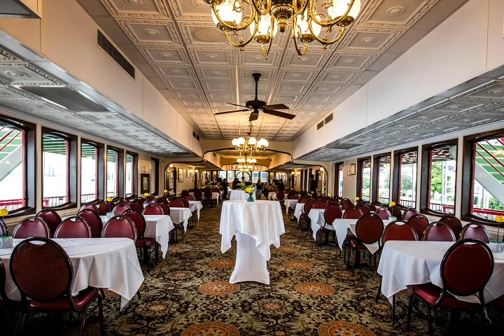 Elegant dining room with patterned carpet, chandeliers, red chairs, and white tablecloths.