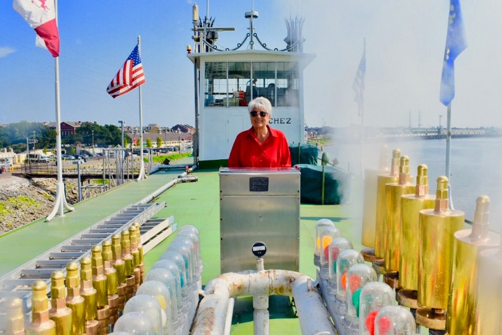 Person in red shirt stands on a ship deck with flags and brass instruments, near water.