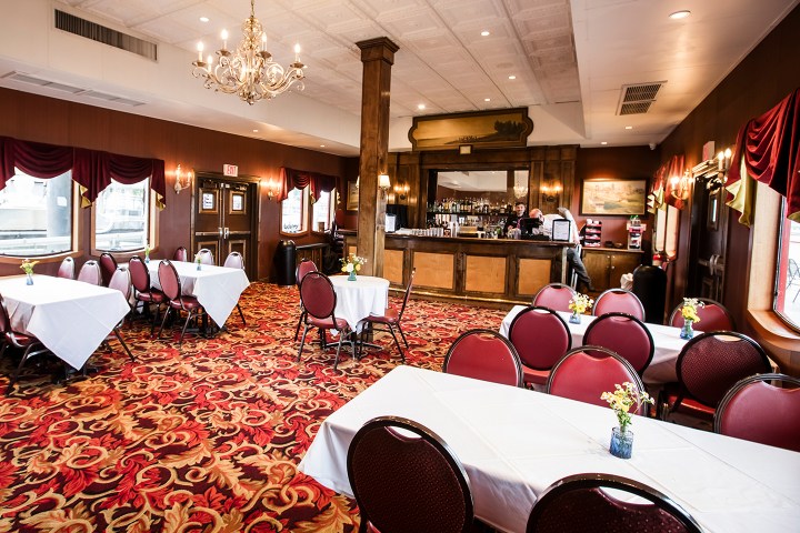 Empty restaurant with red chairs, white tablecloths, and a bar. Chandeliers and floral carpet adorn the space.