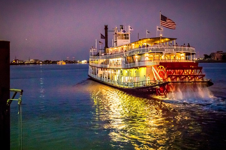Illuminated steamboat on a river at dusk with city lights in the background.