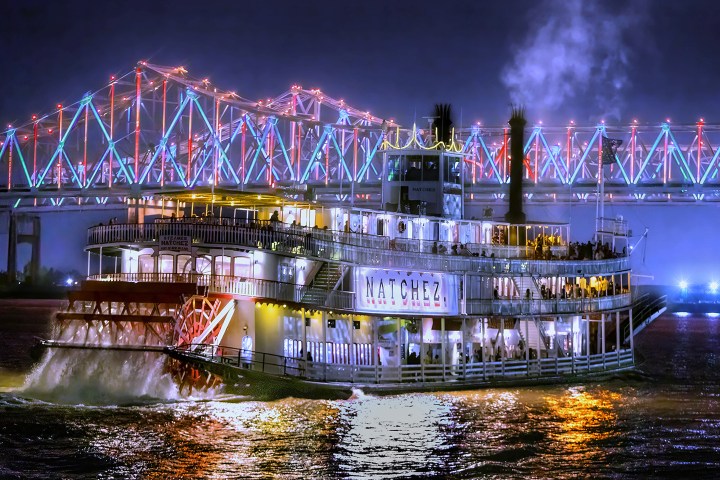 Lit paddle steamer 'Natchez' on river at night, with illuminated bridge in background.