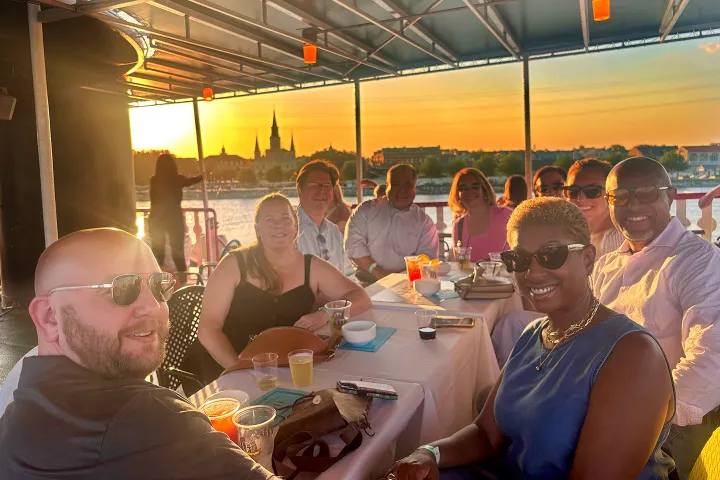 Group of people at a table on a boat during sunset, smiling and enjoying drinks.