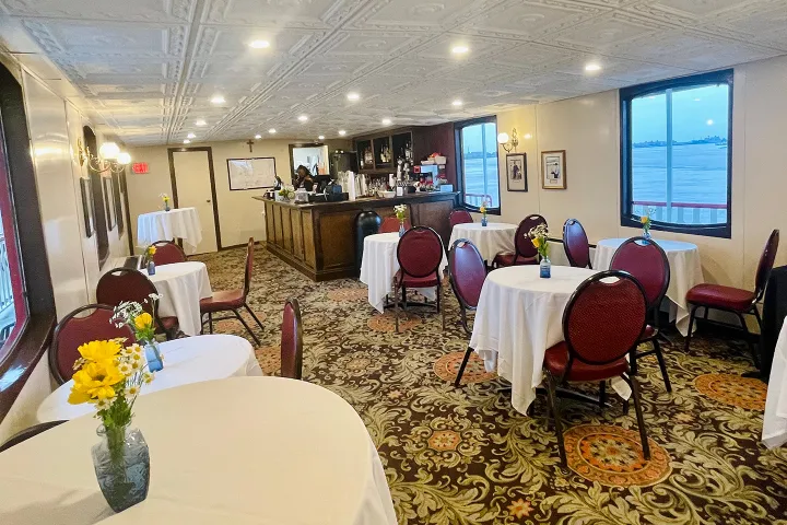 Cozy interior of a boat with round tables, chairs, and a bar, featuring floral arrangements on tables.