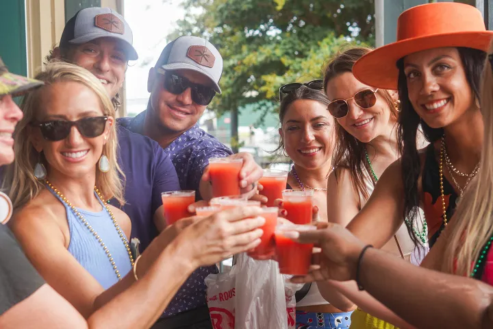Group of people smiling, holding red drinks, wearing hats and sunglasses outdoors.