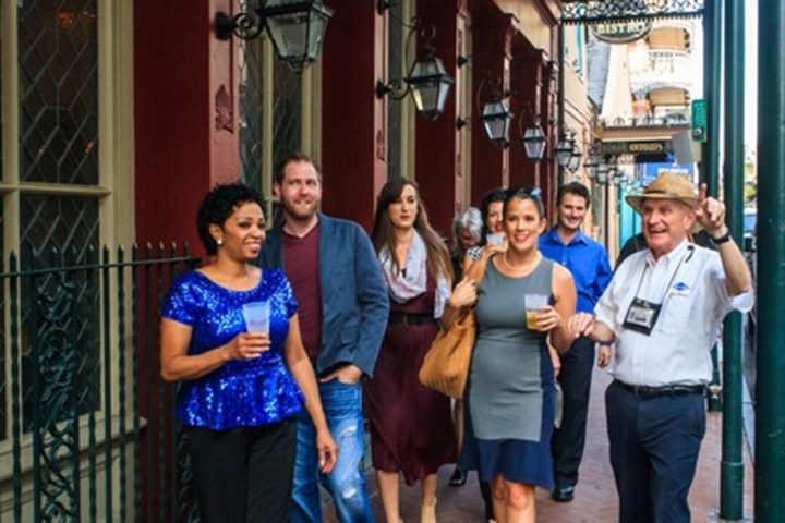 Group of people walking on a lively street, two holding drinks, with a guide gesturing joyfully.
