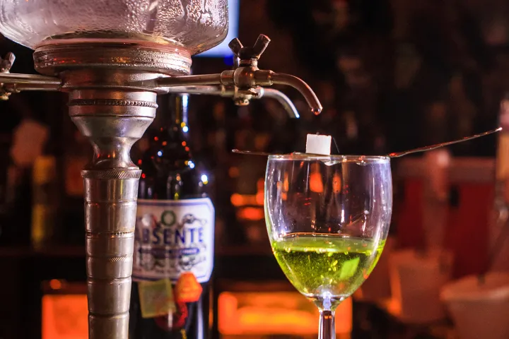 Absinthe being prepared with sugar cube and spoon over a glass under a vintage fountain.