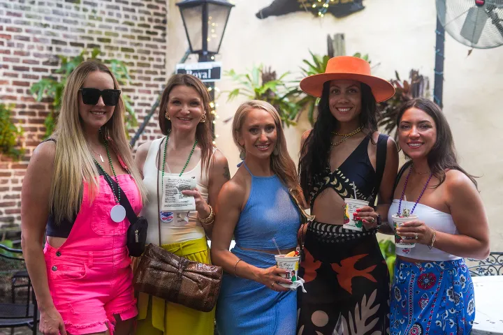 Five women in colorful outfits smiling, holding drinks in an outdoor setting.