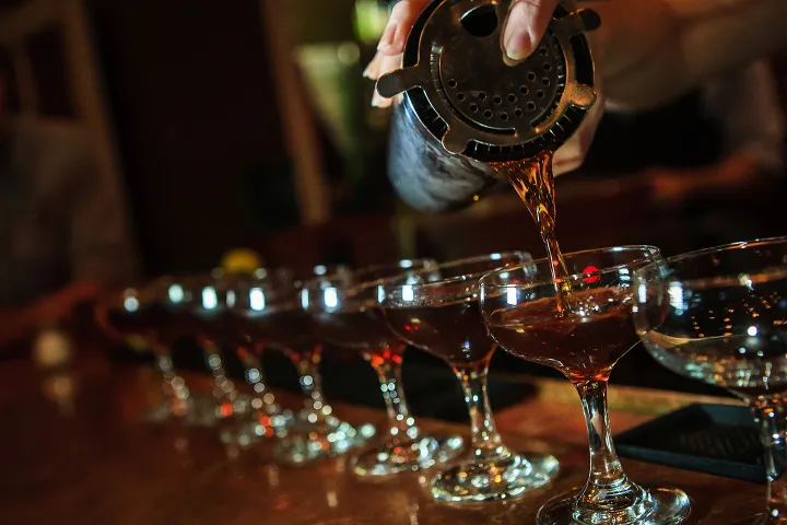Bartender pouring cocktails from shaker into a row of glasses on a bar.