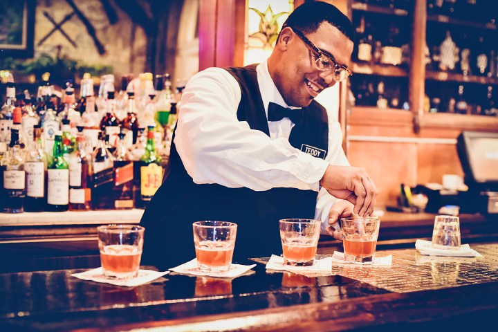 Bartender in white shirt preparing drinks behind a bar with liquor bottles.
