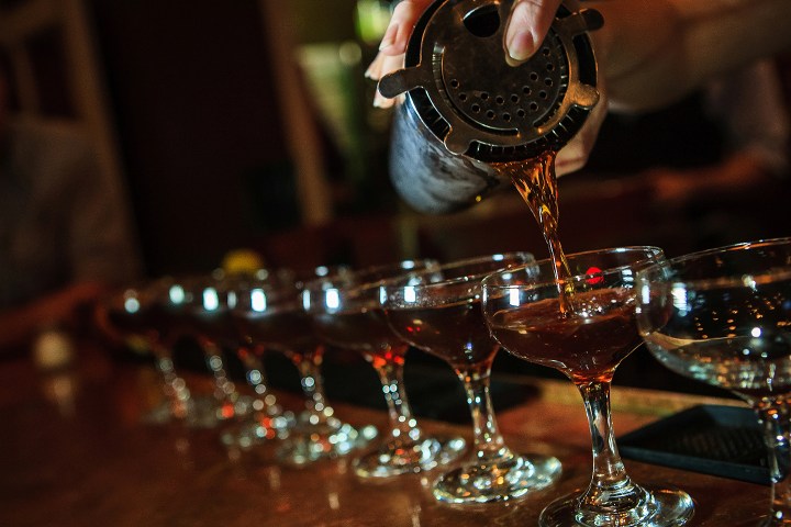 Bartender pouring cocktails into a row of glass cups at a bar.