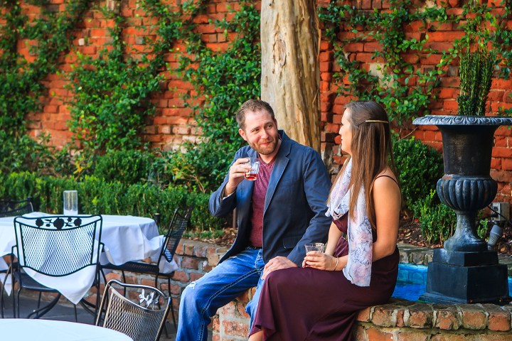 Man and woman sitting outdoors, holding drinks, with brick wall and greenery in the background.