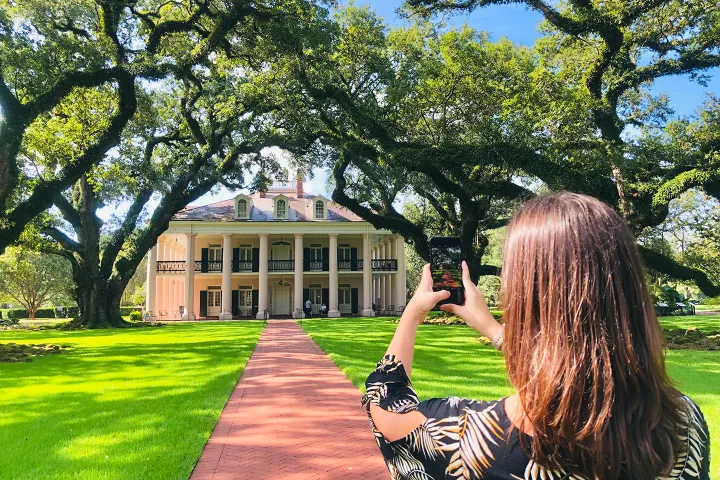Woman photographing plantation-style house with large oak trees on a sunny day.