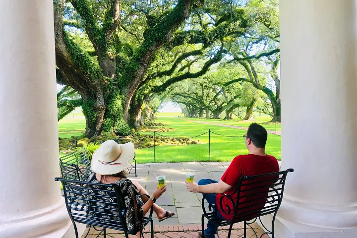 Two people sitting on a porch, facing a garden with large, mossy oak trees.