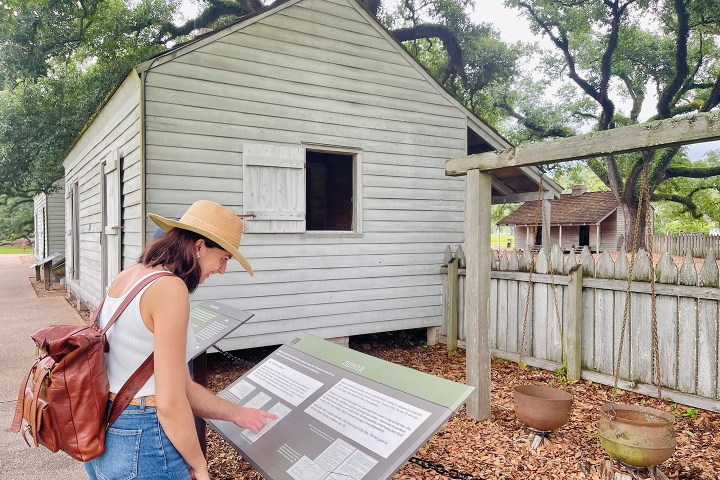 Woman in hat reads plaque near historic wooden house with fences and trees.