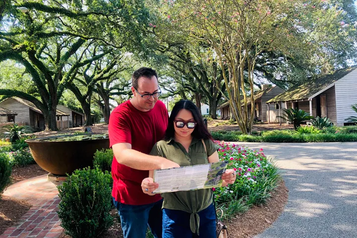 A couple looks at a map in a garden with large trees and historic wooden houses.