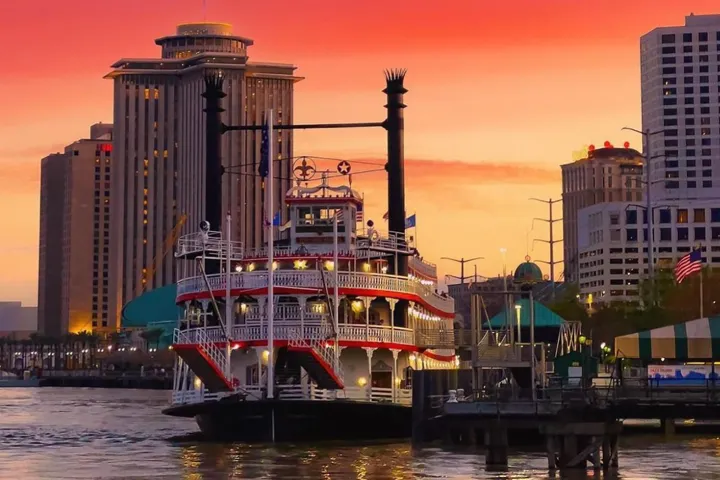 Riverboat on water with city skyline and colorful sunset in background.