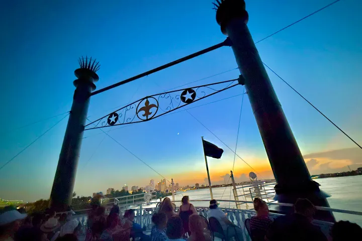 People on a boat deck at sunset, with a city skyline and decorative elements overhead.