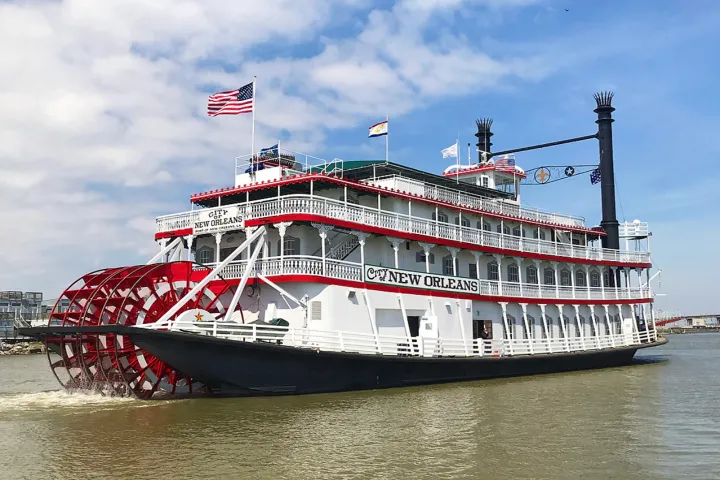 White paddle steamer with red accents on the river, flags waving on top.