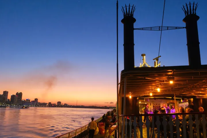 Riverboat with lit deck at sunset, city skyline in background.