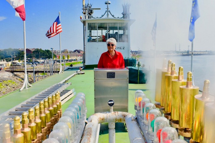Person in red shirt on boat deck with flags and river background.