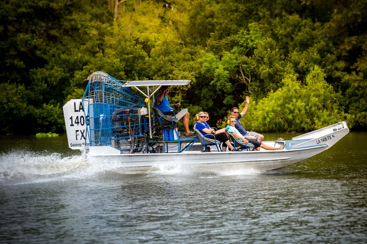 Airboat with five people cruising on a river with lush green trees in the background.