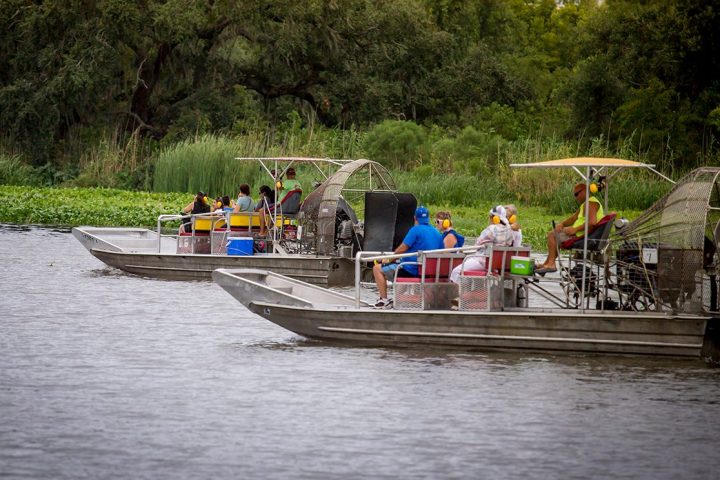 Two airboats with passengers on a river near a forested shoreline.