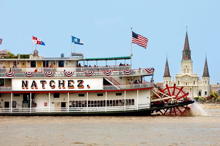 Paddle steamboat Natchez on a river with a historic cathedral in the background.