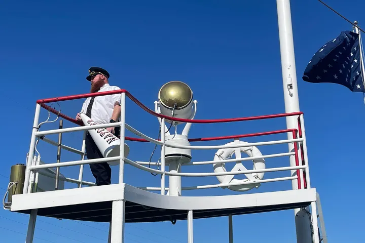 Man in uniform on ship's deck with megaphone and star-spangled flag.