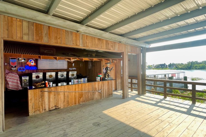 Outdoor wooden bar with drink machines and neon signs, overlooking a sunny waterfront deck.