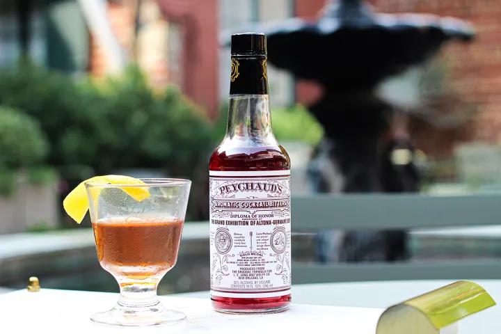 Bottle of bitters beside a cocktail with lemon twist on an outdoor table.
