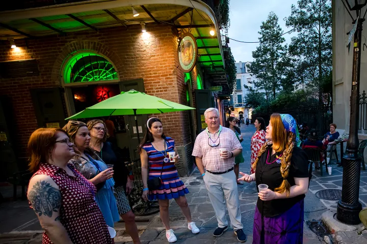 People stand outside a lit brick building, holding drinks, under a green umbrella.