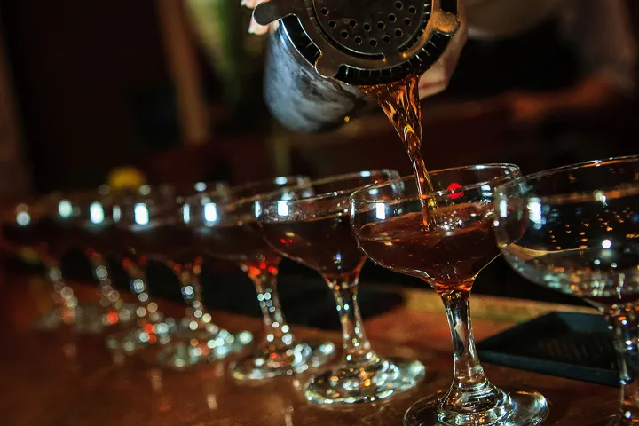 Row of cocktails being poured from a shaker into glasses on a bar.