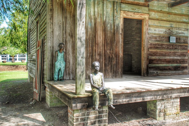 Two child statues sit and stand on a wooden porch of an old cabin.