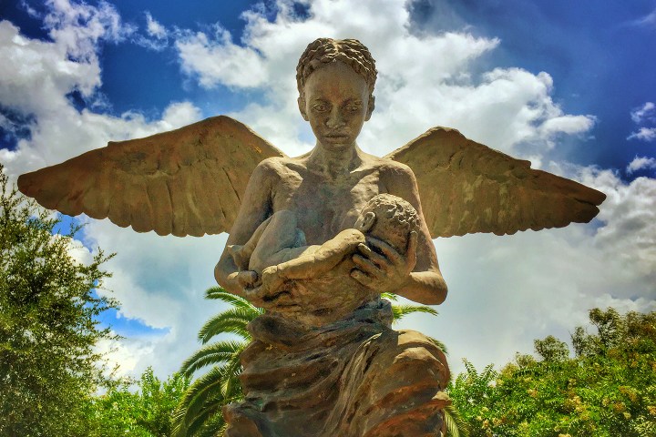 Statue of angel with wings holding a baby against a cloudy sky.