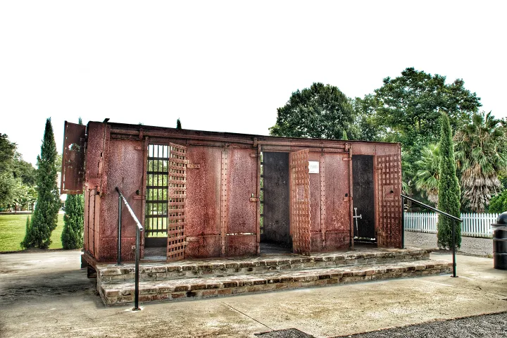 Rusty outdoor jail cell structure on a concrete slab, surrounded by trees.