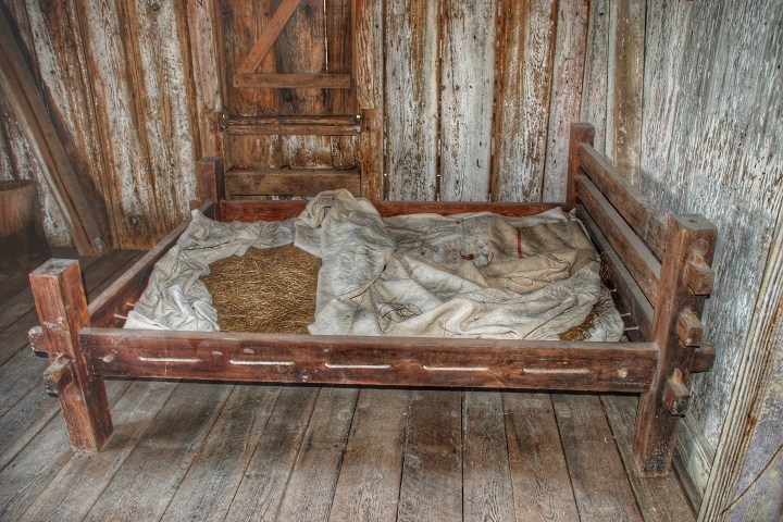 Rustic wooden bed with straw and blankets in a weathered wooden room.