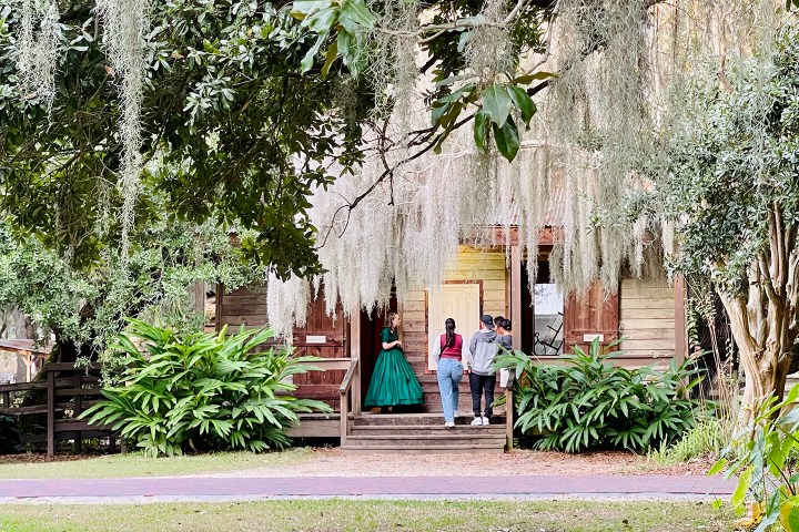 People on porch of rustic house surrounded by greenery and moss-covered trees.