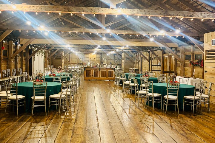 Rustic event hall with round tables, green tablecloths, silver chairs, and string lights.
