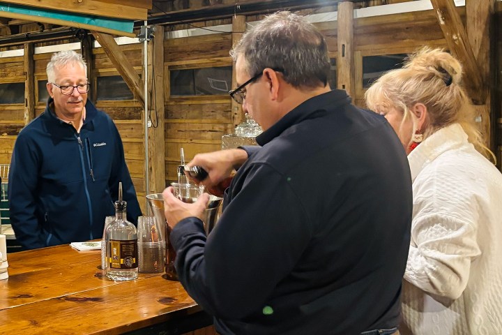 Three people in a wooden bar area; one mixes a drink while the others watch.