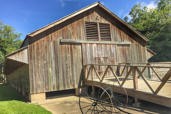 Historic wooden barn with slanted roof and wooden bridge on a sunny day with trees in the background.