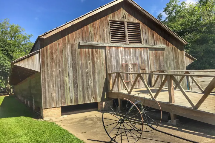 Wooden barn with a ramp and old wagon wheel, surrounded by trees, under a clear blue sky.
