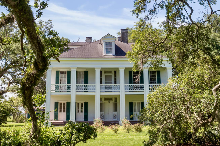 Historic white plantation house with green shutters and columns, surrounded by trees.