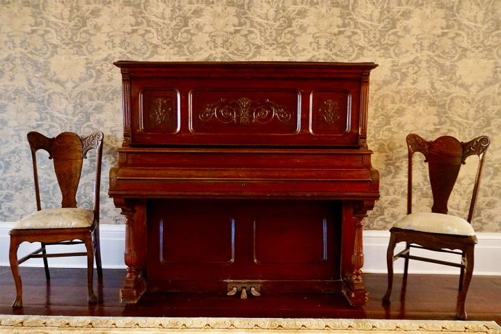Antique upright piano flanked by two ornate wooden chairs on a wooden floor.